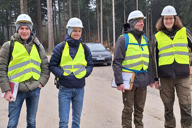 Studierende mit neongelben Warnwesten und Helmen stehen als Gruppe auf der Baustelle  im Hoßkircher Wald