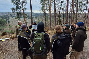 Studierende stehen in einem Halbkreis auf einem Waldweg