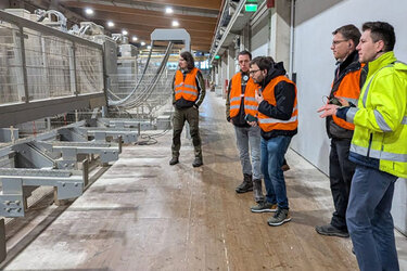 Studierende stehen mit Warnwesten im Sägewerksgebäude und betrachten eine Maschine.